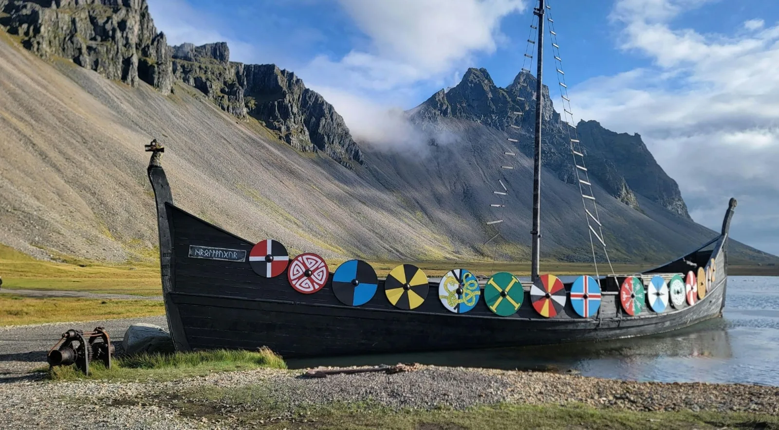 A Viking longship replica with painted shields lined along its hull, beached beside a fjord with steep mountains behind