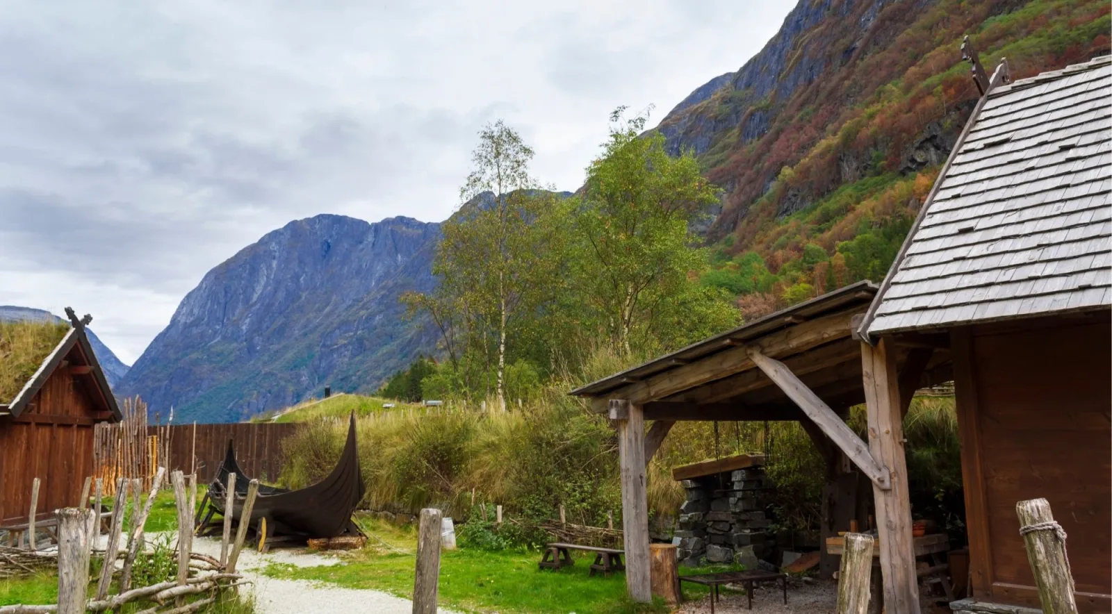 A cluster of turf-roofed wooden buildings with a small Viking boat in the yard, mountains rising behind