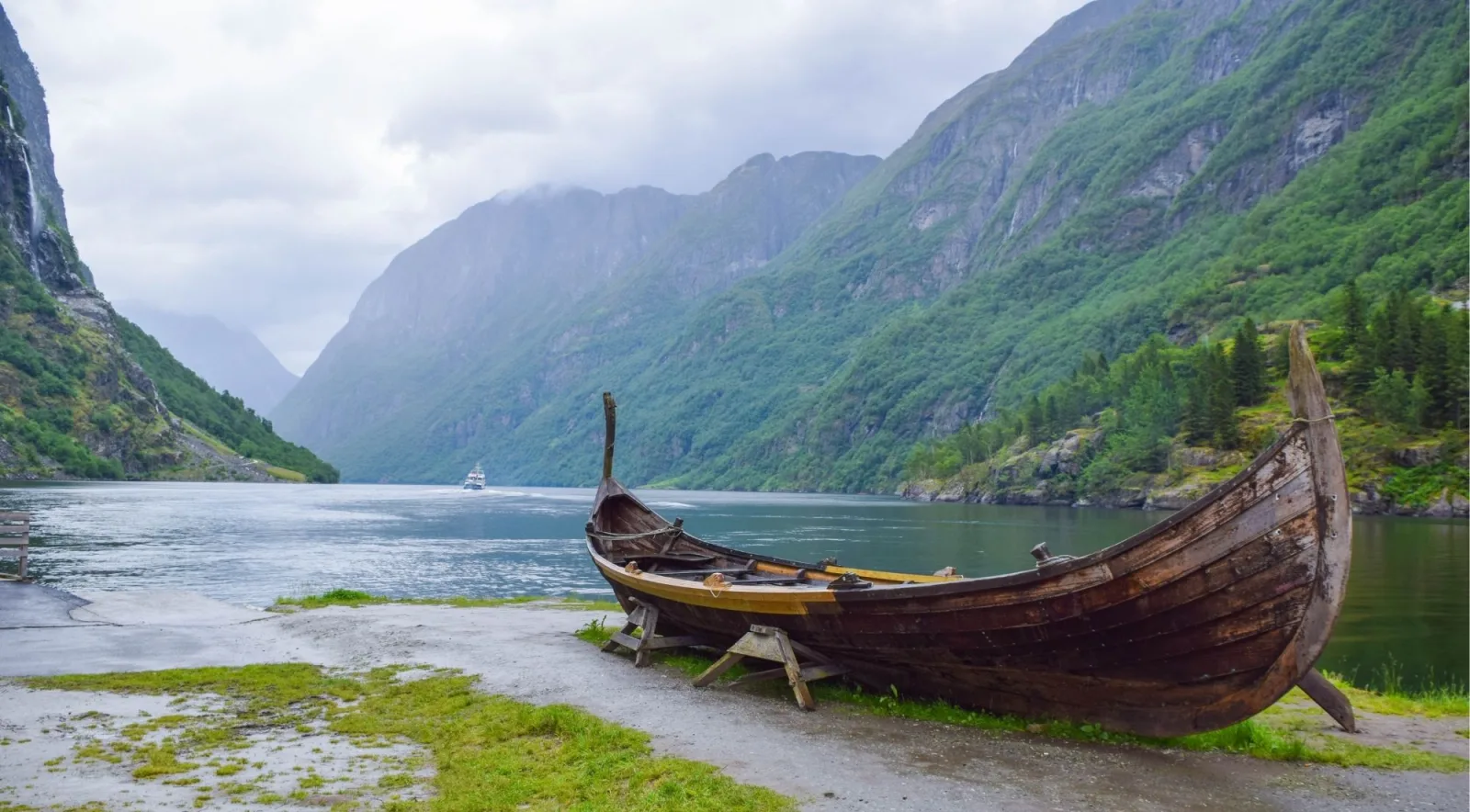 A wooden Viking longboat on the shore of a narrow fjord, with a cruise vessel passing in the distance