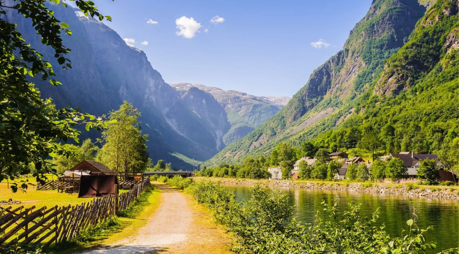 A gravel path beside a fjord leading toward a small village of white and red houses, mountains behind